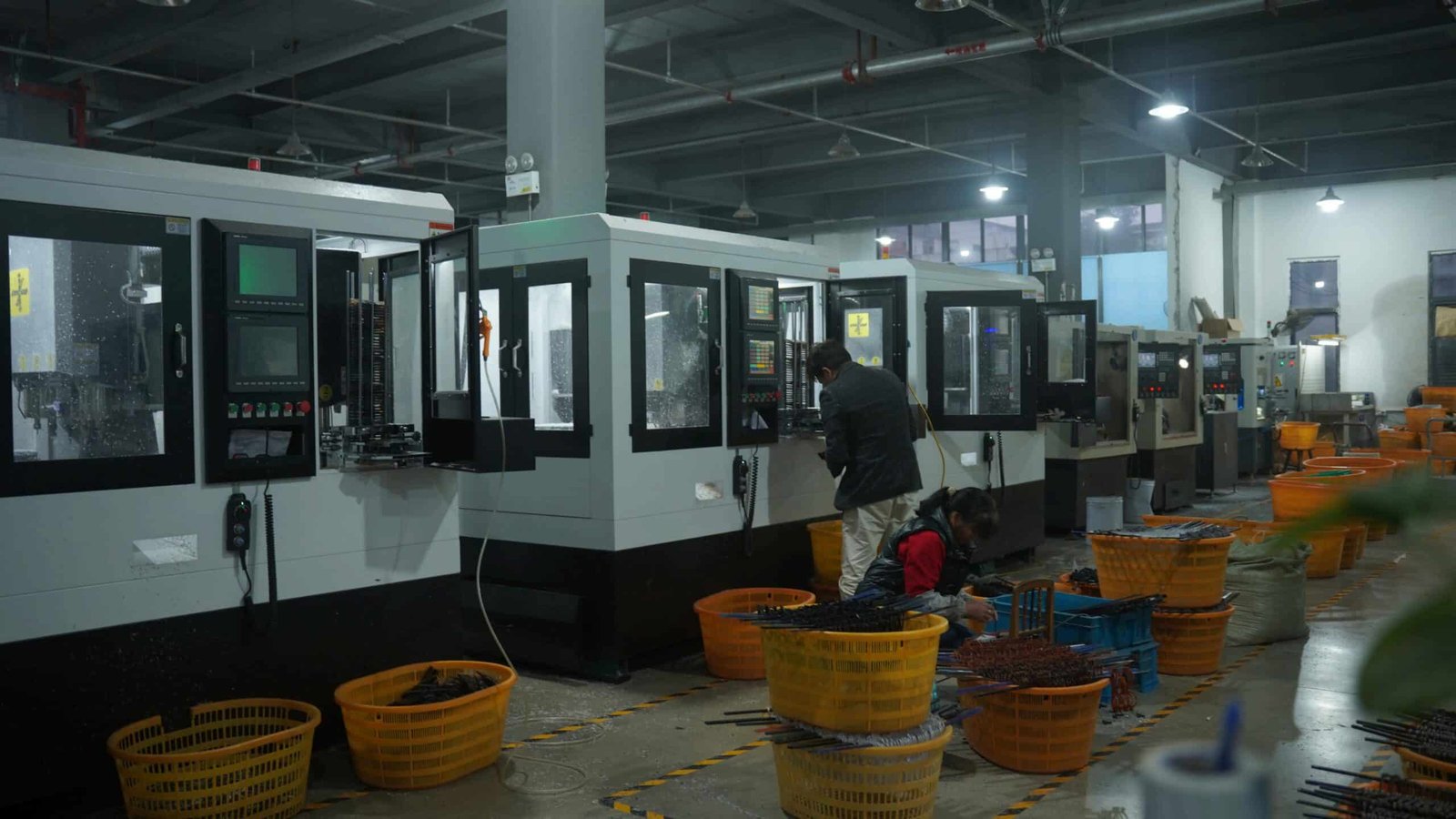 A factory floor with several large white CNC machines. A male worker is standing and operating a machine's control panel, while a female worker sits nearby, sorting long, dark manufactured components into orange plastic baskets. Multiple orange baskets filled with parts are scattered across the marked concrete floor.
